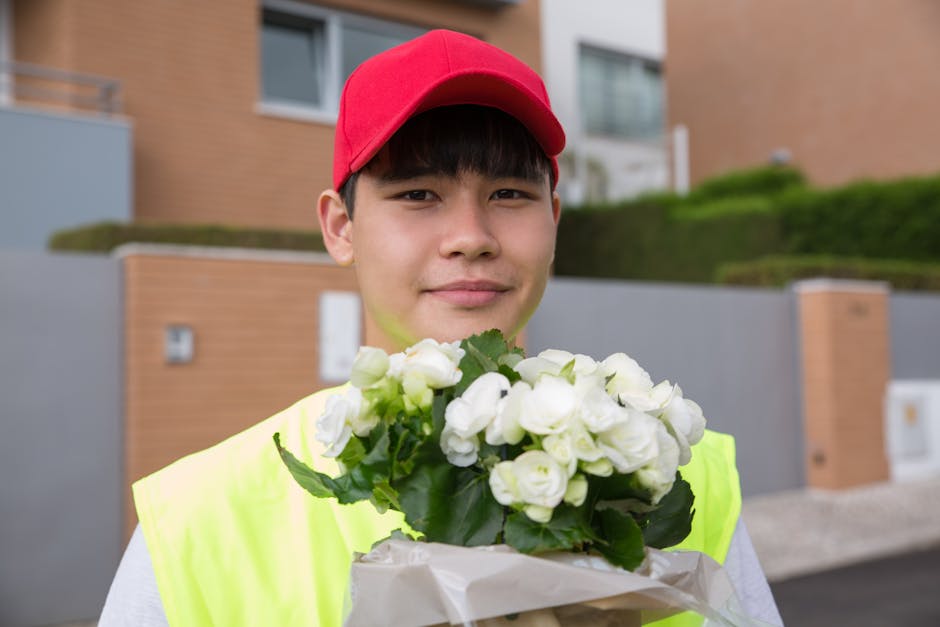 A young male delivery person wearing a red cap and a high-visibility yellow vest is holding a bouquet of white flowers wrapped in white paper. He is standing outdoors in front of a modern residential