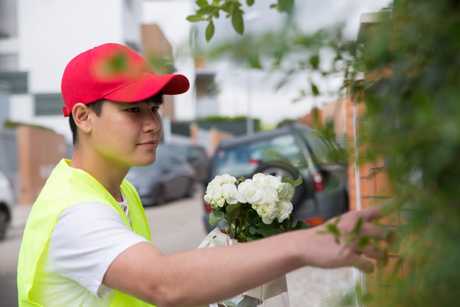 A young male florist wearing a red cap and a neon yellow vest is seen delivering a floral gift outdoors. He holds a bouquet of fresh white roses with green leaves, wrapped in white paper. The arrangem