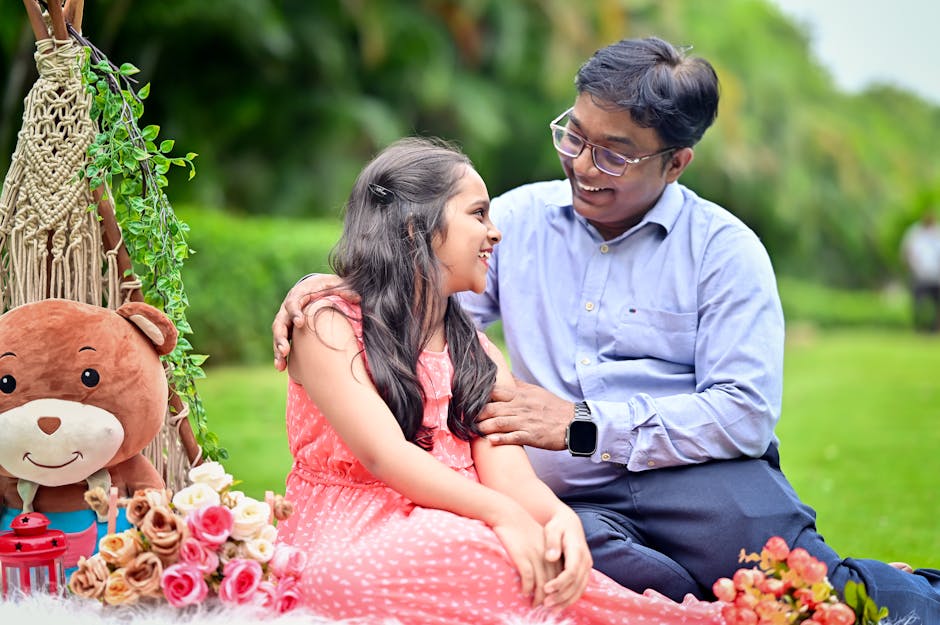 A young girl with long dark hair wearing a pink sleeveless dress is sitting outdoors on green grass, smiling joyfully as an older man, dressed in a light blue shirt and dark trousers, gently touches h