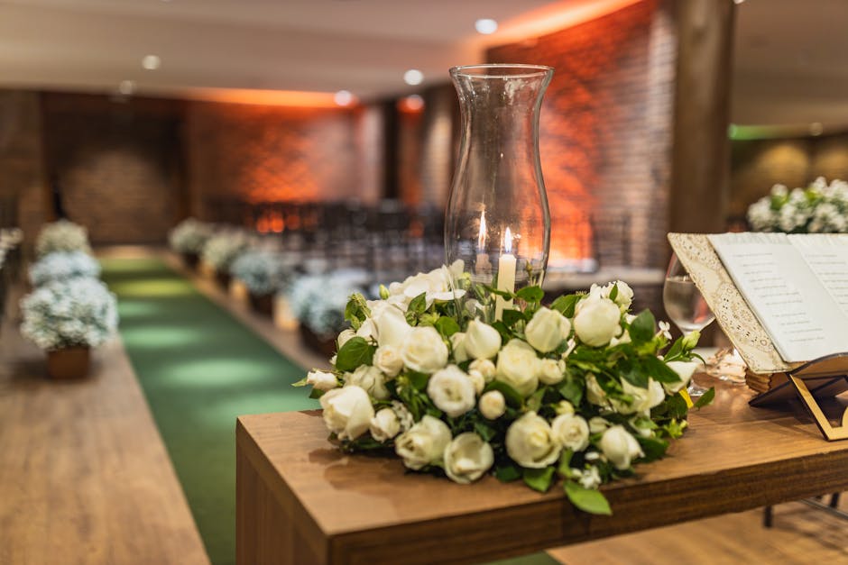 A floral arrangement featuring a bouquet of small white roses and white lisianthus, accented with green foliage, is placed on a wooden table. The bouquet surrounds a tall, clear glass hurricane lanter