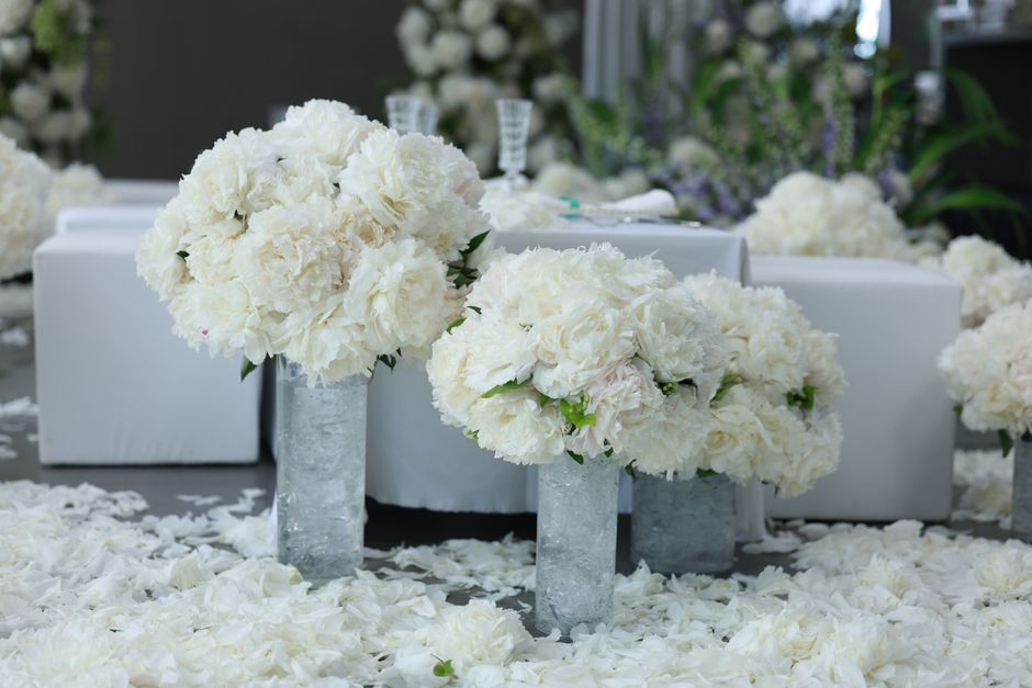 A floral arrangement featuring large, round bouquets of white peonies and roses, arranged in tall, textured glass vases. The flowers appear fresh with soft, delicate petals and sturdy green stems visi