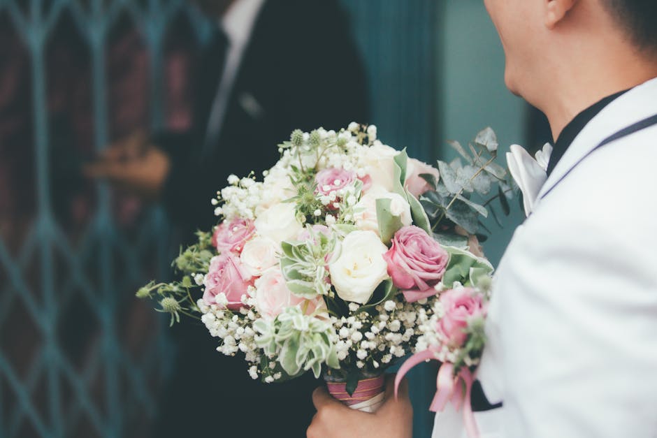 A man dressed in formal attire, holding a floral bouquet arrangement consisting of white and pink roses, baby's breath, and greenery, wrapped with pink ribbon. The bouquet features fresh, tightly peta