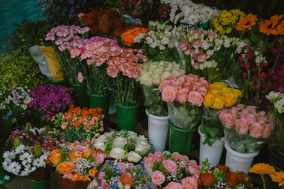 A colorful display of various fresh flower bouquets arranged in white and green vases and wrapped in clear plastic, featuring roses in shades of pink, yellow, and white, as well as carnations, daisies